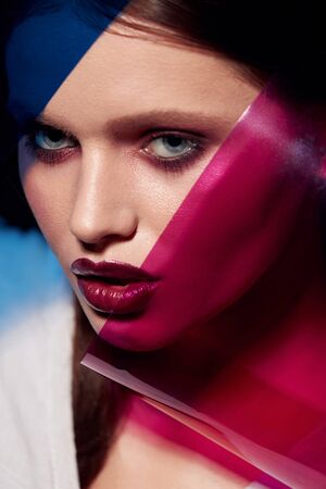Beauty face makeup. Fashion portrait of girl model with lipstick under color. Closeup of glamorous girl with sexy dark red lips make-up and glowing skin under pink and blue colors. High qualityの写真素材