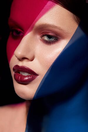 Beauty face makeup. Fashion portrait of girl model with lipstick under color. Closeup of glamorous girl with sexy dark red lips make-up and glowing skin under pink and blue colors. High qualityの写真素材