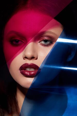 Beauty face makeup. Fashion portrait of girl model with lipstick under color. Closeup of glamorous girl with sexy dark red lips make-up and glowing skin under pink and blue colors. High qualityの写真素材