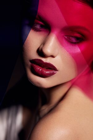 Beauty face makeup. Fashion portrait of girl model with lipstick under color. Closeup of glamorous girl with sexy dark red lips make-up and glowing skin under pink and purple colors. High qualityの写真素材