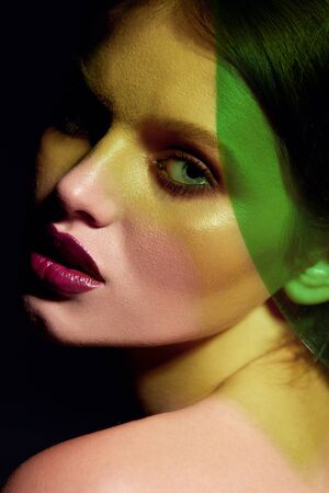 Fashion model with beauty face makeup under green neon color. Closeup portrait of woman with glamour make up look, dark lipstick on lips and facial skin glow under colorful light. High qualityの写真素材
