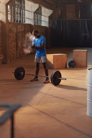 Sport man using hand chalk powder before workout at gym. Black male athlete applying magnesium carbonate gym chalk before lifting barbell, exercising at fitness club indoorsの写真素材