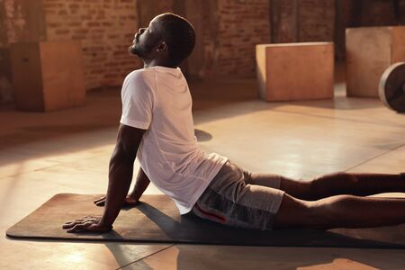Body stretch. Fit man stretching back, doing yoga workout at gym. Black male athlete doing back extension stretch exercise lying on mat indoorsの写真素材