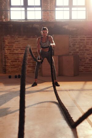Rope workout. Sport woman doing battle ropes exercise at gym. Fitness girl athlete with fit body exercising, doing functional training with heavy ropes indoorsの写真素材