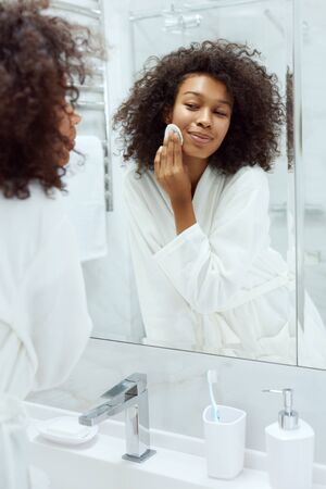 Face skin care. Girl removing makeup with cosmetic cotton pad at bathroom. Closeup portrait of beautiful smiling african woman with afro hair cleaning facial skin with white padの写真素材