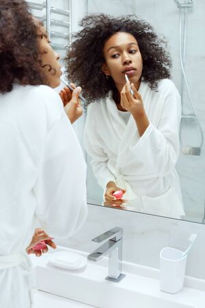 Lips skin care. Woman applying lip balm with makeup brush looking in mirror at bathroom. Portrait of beautiful african girl model with beauty face applying lip productの写真素材