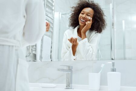 Skin care. Woman applying cosmetic cream on face looking in mirror at bathroom. Portrait of smiling female model with natural makeup, clean facial skin applying moisturizing lotionの写真素材
