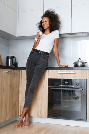 Drink water. Happy young woman with glass of fresh water in white kitchen portrait. Beautiful smiling african american girl drinking pure mineral water in morning at homeの写真素材