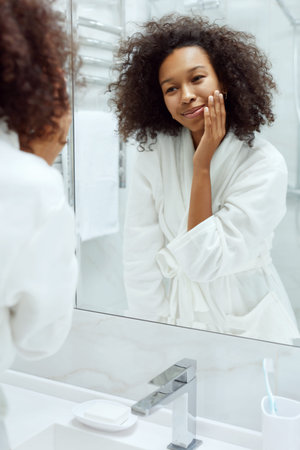 Skin care. Smiling woman touching face skin, looking at mirror at bathroom. Portrait of beautiful...