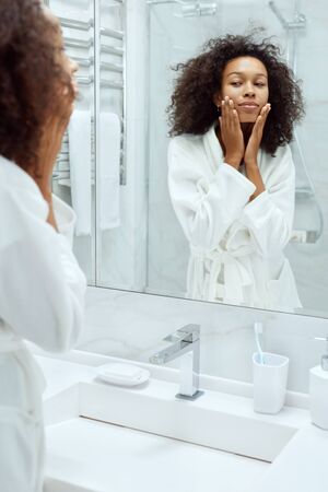 Skin care. Woman touching face skin and looking at mirror at bathroom. Portrait of beautiful african girl with afro hair after shower going through her morning beauty routineの写真素材