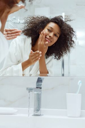Skin care. Woman washing face with clean water in sink looking in mirror at bathroom. Portrait of beautiful smiling african girl with afro hair in white bath robe cleaning facial skin closeupの写真素材