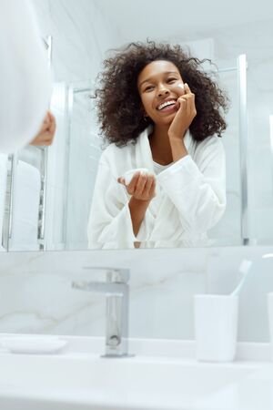 Skin care. Woman applying cosmetic cream on face looking in mirror at bathroom. Portrait of smiling female model with natural makeup, clean facial skin applying moisturizing lotionの写真素材