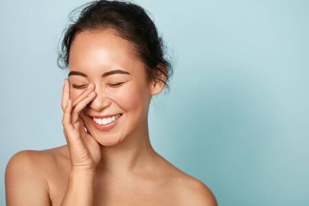 Beauty face. Smiling asian woman touching healthy skin portrait. Beautiful happy girl model with fresh glowing hydrated facial skin and natural makeup on blue background at studio. Skin care concept