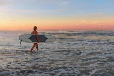 Surfer. Surfing Man With White Surfboard Walking In Ocean. Water Sport For Active Lifestyle. Beautiful Tropical Sunset At Sea.の写真素材