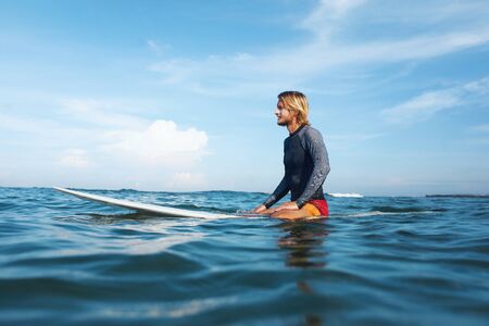 Surfing. Surfer Man Sitting On Surfboard Portrait. Handsome Guy In Wetsuit In Ocean. Blue Sea And Beautiful Sky With Soft Clouds On Background.の写真素材