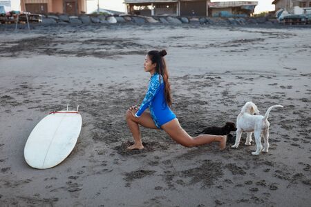 Surfer Girl. Surfing Woman Stretching On Sandy Beach. Asian Brunette In Blue Wetsuit With Surfboard And Dog. Water Sport For Active Lifestyle.の写真素材