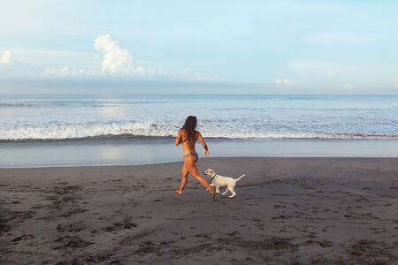 Sexy Girl. Tanned Woman Running On Sandy Beach. Beautiful Brunette In Bikini Playing With Dog Near Ocean.の写真素材