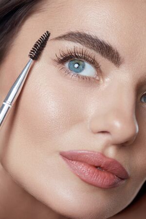 Makeup. Woman Brushing Eyebrows With Brush Close Up Portrait. Beautiful Brunette With Perfect Skin. Blue-Eyed Girl Looking Up.の写真素材