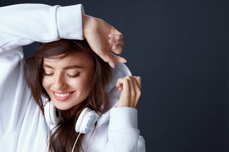 Stylish Girl. Beautiful Model Close Up Portrait. Smiling Woman With Headphones On Neck Holding Hands On Head. Pretty Brunette With Closed Eyes In White Hoodie On Black Background.の写真素材