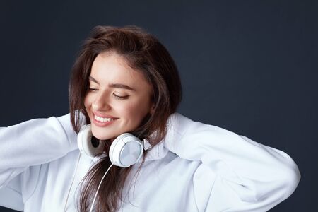 Stylish Girl. Beautiful Woman Close Up Portrait. Smiling Model With Headphones On Neck Holding Hands Behind Head. Pretty Brunette In White Hoodie On Black Background.の写真素材