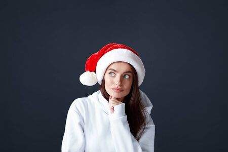 Santa Girl. Beautiful Woman In Christmas Hat Portrait. Thinking Model Keeps Hand On Chin. Brunette In White Hoodie On Black Background. Blue-Eyed Female Looking Away.の写真素材