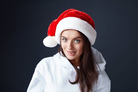 Santa Girl. Beautiful Woman In Christmas Hat Portrait. Model Biting Lips And Looking At Camera. Brunette In White Hoodie On Black Background.の写真素材