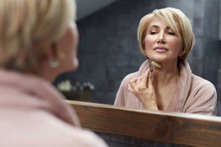 Skincare Routine. Mature Woman Uses Jade Roller On Face Skin In Front Of The Mirror Reflection. Anti Aging Beauty Treatment Concept.の写真素材