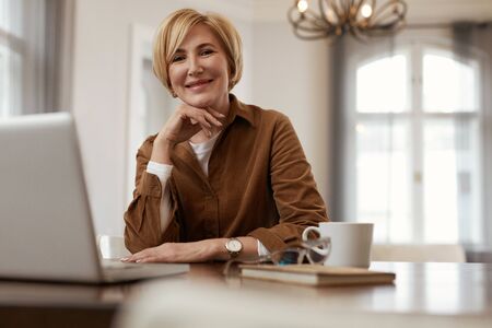 Female With Laptop. Senior Blonde Woman In Brown Jacket Sits At The Desk With Laptop And Cup. Businesswoman Homework Concept.の写真素材
