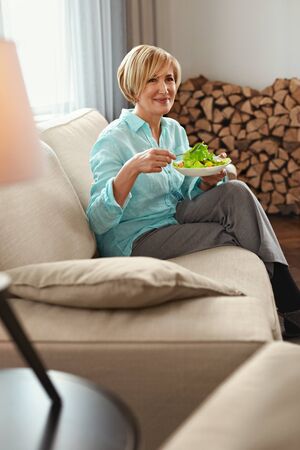 Mature Woman Eats Salad. Middle Age Female In Casual Clothes With Healthy Food On Couch At Home. Concept of Nutrition.の写真素材