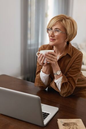 Businesswoman. Mature Woman In Brown Jacket And Eyeglasses Drinks From Cup. Laptop At The Desk. Remote Homework Concept.の写真素材