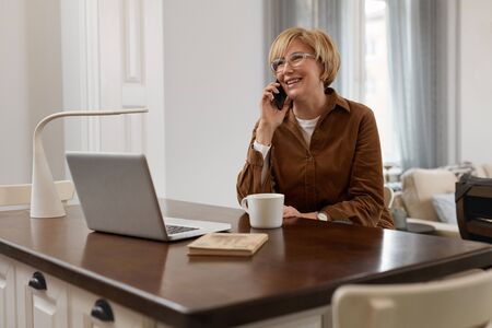 Businesswoman Homework. Female With Laptop. Mature Woman In Brown Jacket And Eyeglasses Talks On Phone. Laptop At The Desk.の写真素材