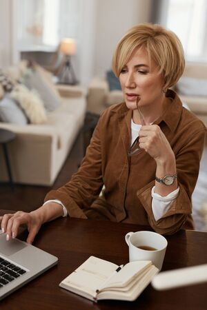 Businesswoman. Female With Laptop. Mature Woman Remote Homework Concept. Model in Brown Jacket And Eyeglasses Uses Notebook.の写真素材