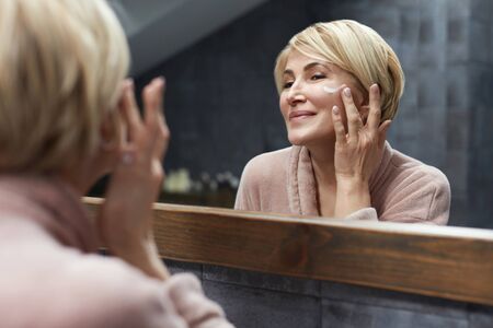 Skincare Routine. Mature Woman Uses Cosmetic Cream On Face Skin In Front Of The Mirror Reflection. Anti Aging Beauty Treatment Concept.の写真素材