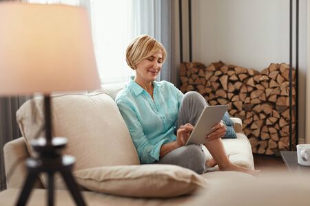 Female With Tablet. Mature Woman On Sofa In Beige Plaid Uses Digital Device Touchscreen, Comfortable Internet Search. High Resolution.の写真素材
