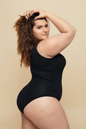 Plus Size Model. Fat Woman In Black Bodysuit Portrait. Brunette Touching Hair And Looking At Camera. Body Positive Concept On Beige Background.の写真素材