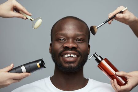 Male Skin Care Routine. Facial Beauty Treatment For African Model. Jade Roller, Electric Razor, Makeup Brush And Liquid In Bottle Near Man's Face.の写真素材