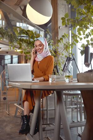 Muslim Woman. Young Girl In Hijab Portrait. Beautiful Female Sitting In Cafe And Working On Laptop. Smiling Model Looking At Screen And Talking.の写真素材