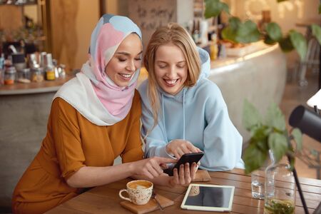 Friends. Young Girls In Cafe. Smiling Women Looking At Smartphone Screen. Different Ethnicity Female Spending Time In Bistro.の写真素材
