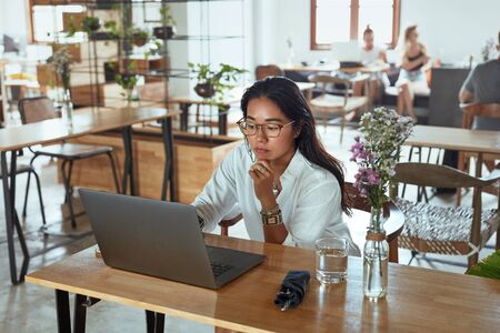 Business. Woman Working Remotely At Cafe Portrait. Girl In Glasses Using Laptop And Waiting For Lunch In Restaurant. Model Touching Chin And Looking At Screen.の写真素材