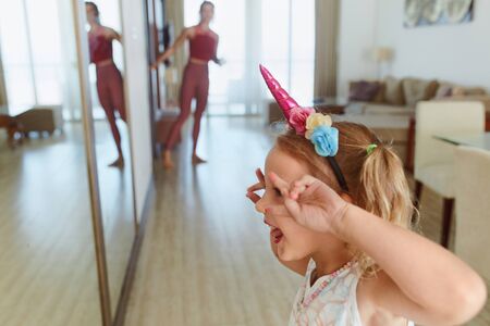 Family. Child And Mother Having Fun At Home. Little Girl Standing In Front Of Mirror And Making Funny Face. Young Woman In Sportswear Looking For Kid From Another Room.の写真素材
