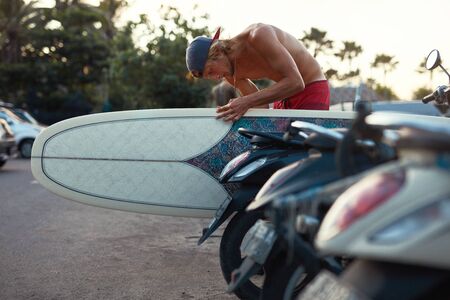 Surfing. Handsome Surfer Waxing White Surfboard On Beach Road. Young Guy In Cap At Sunset. Water Sport For Active Lifestyle.の写真素材