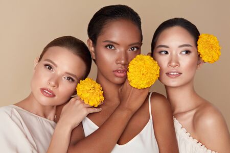 Beauty. Models With Flowers Portrait. Group Of Diversity Women Holding Yellow Marigolds And Posing Against Beige Background. Asian, Mixed Race And Caucasian Female With Natural Makeup.の写真素材