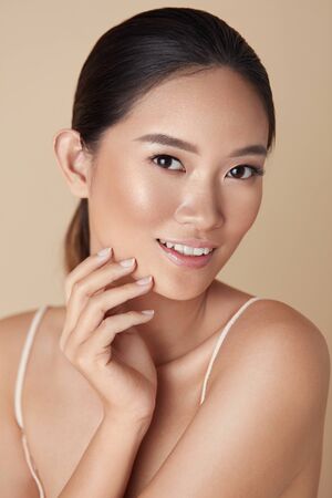 Beauty. Asian Woman Portrait. Beautiful Model Touching Face And Looking At Camera. Charming Girl With Healthy Glowing Skin And Nude Makeup Posing Against Beige Background.の写真素材