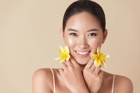 Beauty Face. Flowers And Model Portrait. Asian Woman Holding Plumeria And Looking At Camera Against Beige Background. Female With Natural Nude Makeup And Clean Healthy Skin.の写真素材