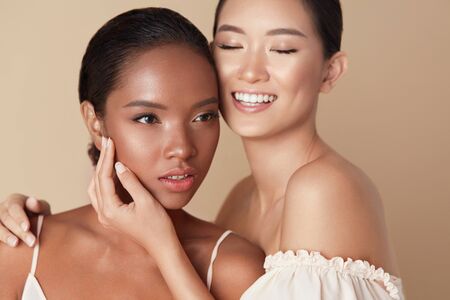 Women. Diversity Models Beauty Portrait. Two Ethnic Female With Nude Makeup And Smooth Hydrated Skin. Smiling Asian Girl Hugs Mixed Race Model And Touches Her Face Against Beige Background.の写真素材