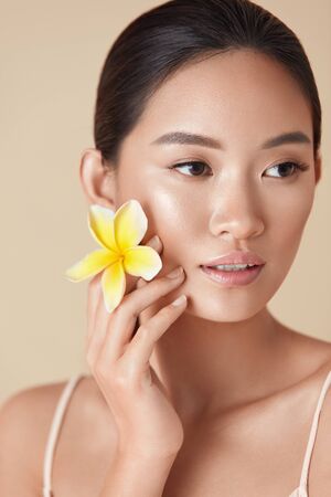 Beauty Face. Flower And Model Close Up Portrait. Beautiful Asian Woman With Plumeria Looking Away Against Beige Background. Girl With Natural Nude Makeup And Clean Healthy Skin After SPA Treatment.の写真素材