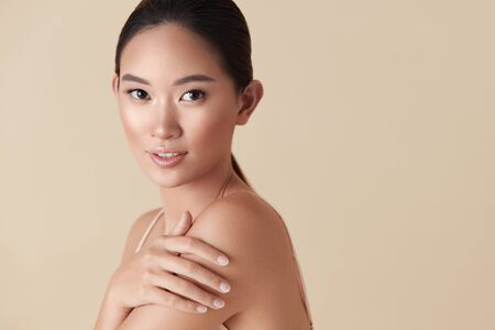 Woman. Asian Model Beauty Portrait. Beautiful Female Touches Her Shoulder And Looking At Camera. Tender Girl With Perfect Glowing Skin And Nude Natural Makeup Posing Against Beige Background.の写真素材