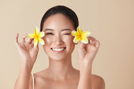 Beauty. Model With Flowers Portrait. Happy Asian Girl With Tropical Plumeria Near Face Against Beige Background. Ethnic Woman With Nude Makeup, Healthy, Smooth And Glowing Facial Skin.の写真素材