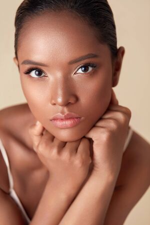 Woman. Ethnic Model Beauty Portrait. Beautiful Mixed Race Female With Hands Under Chin Looking At Camera. Charming Girl With Perfect Skin And Nude Makeup On Beige Background.の写真素材