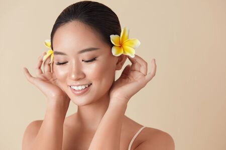 Face. Woman With Flowers Portrait. Beautiful Asian Model With Plumeria In Hair Posing Against Beige Background. Charming Girl With Closed Eyes, Nude Makeup, Healthy, Hydrated, Smooth And Glowing Skin.の写真素材